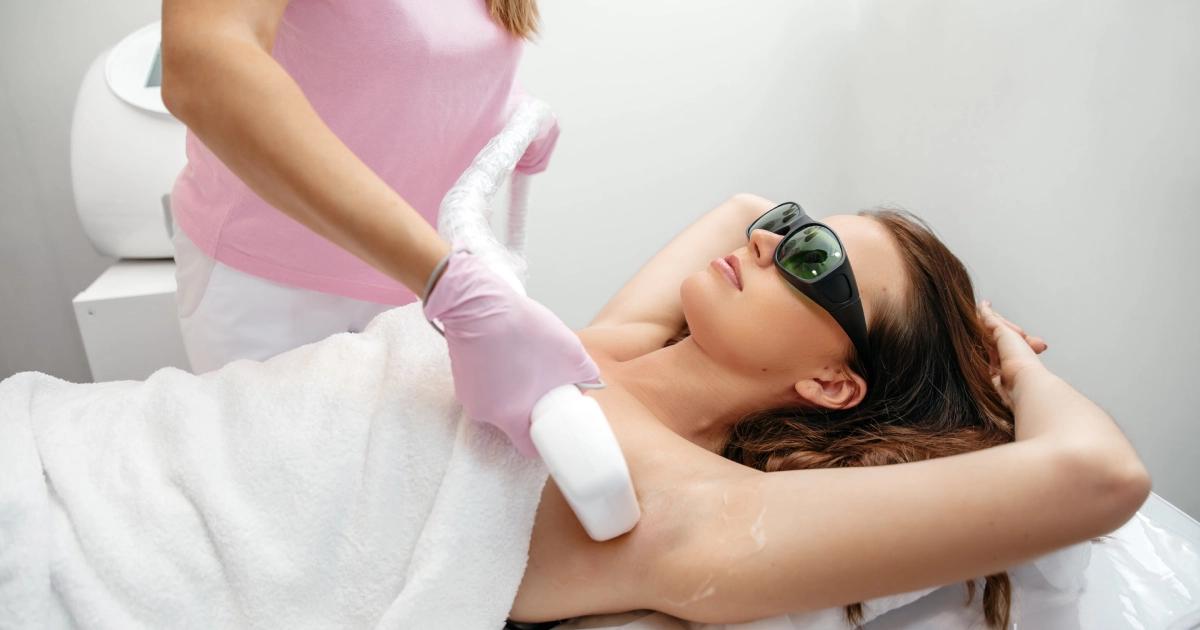 Woman receiving laser hair reduction treatment on underarm by a professional wearing gloves, both using protective eyewear in a clinic setting.